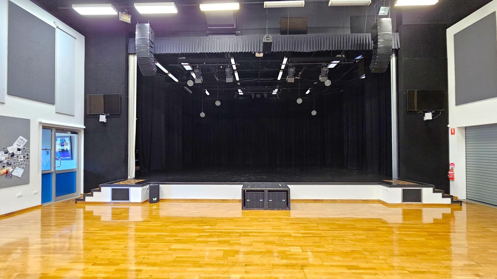 A spacious auditorium with a wooden floor and a black stage at the centre. The stage is equipped with overhead lighting and speakers. On the left, there are noticeboards and a glass door with a blue frame. On the right, a grey roller door and a fire extinguisher are visible. The ceiling is fitted with rectangular lights, and the walls are adorned with acoustic panels.