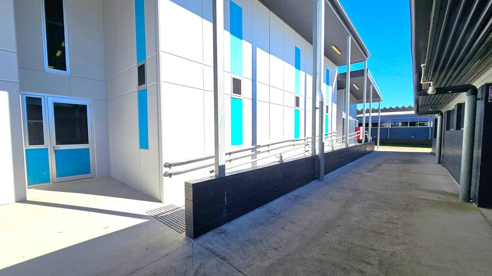 A narrow outdoor corridor with a concrete floor. On the left, a building with white walls and vertical blue panels features a door and windows. A metal railing runs alongside a ramp. On the right, a dark-coloured building with a corrugated roof and small windows. The ceiling is not visible. In the background, another building with a similar design is partially visible under a clear blue sky.