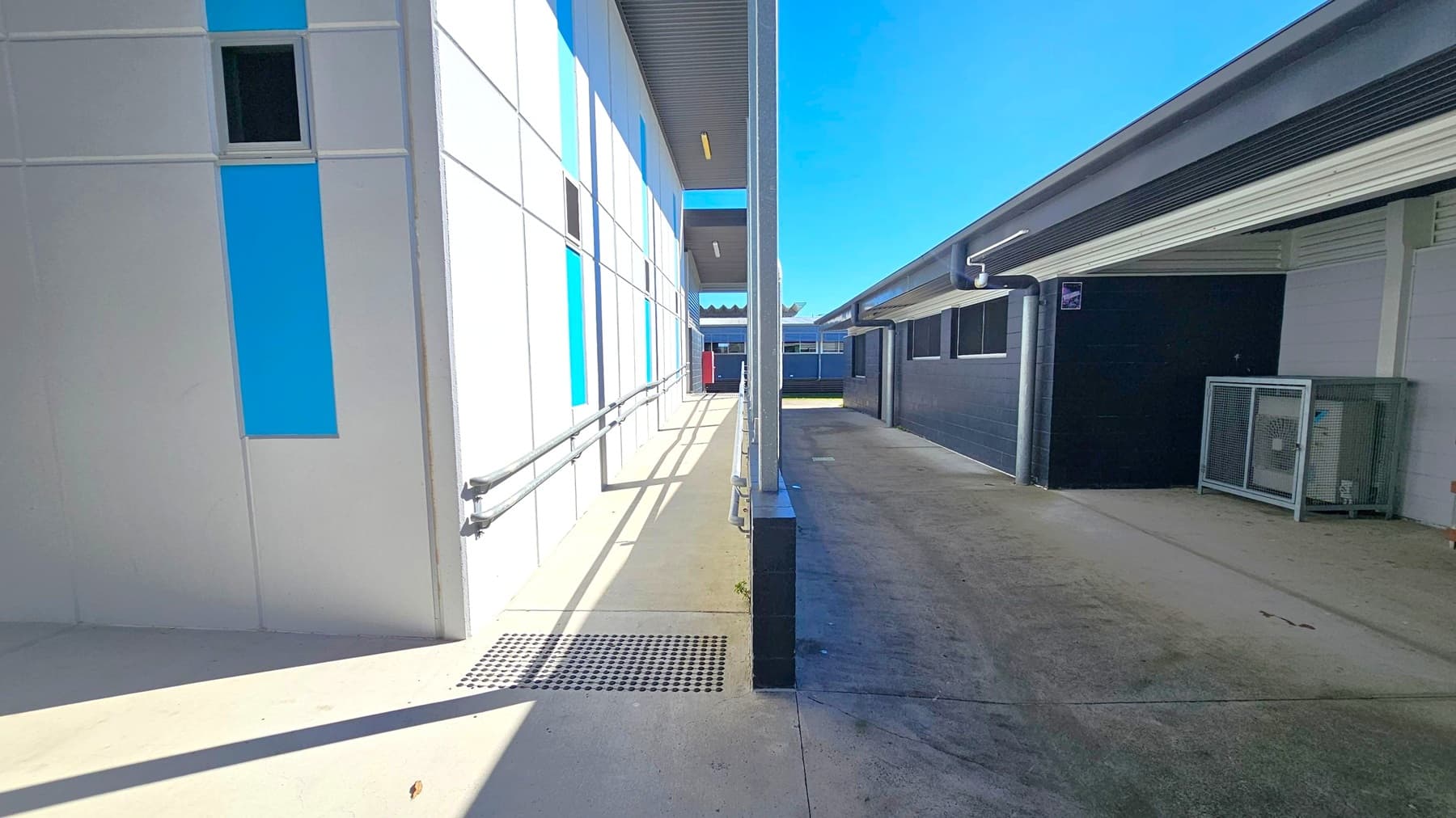 A narrow outdoor corridor with a building on each side. On the left, a white building features vertical blue panels and a small window. A metal handrail runs along the walkway. On the right, a darker building has a series of windows and a covered area with a metal cage structure. The ground is concrete, and the ceiling of the left building has a metal finish with fluorescent lights. The corridor leads to a more open area in the distance.