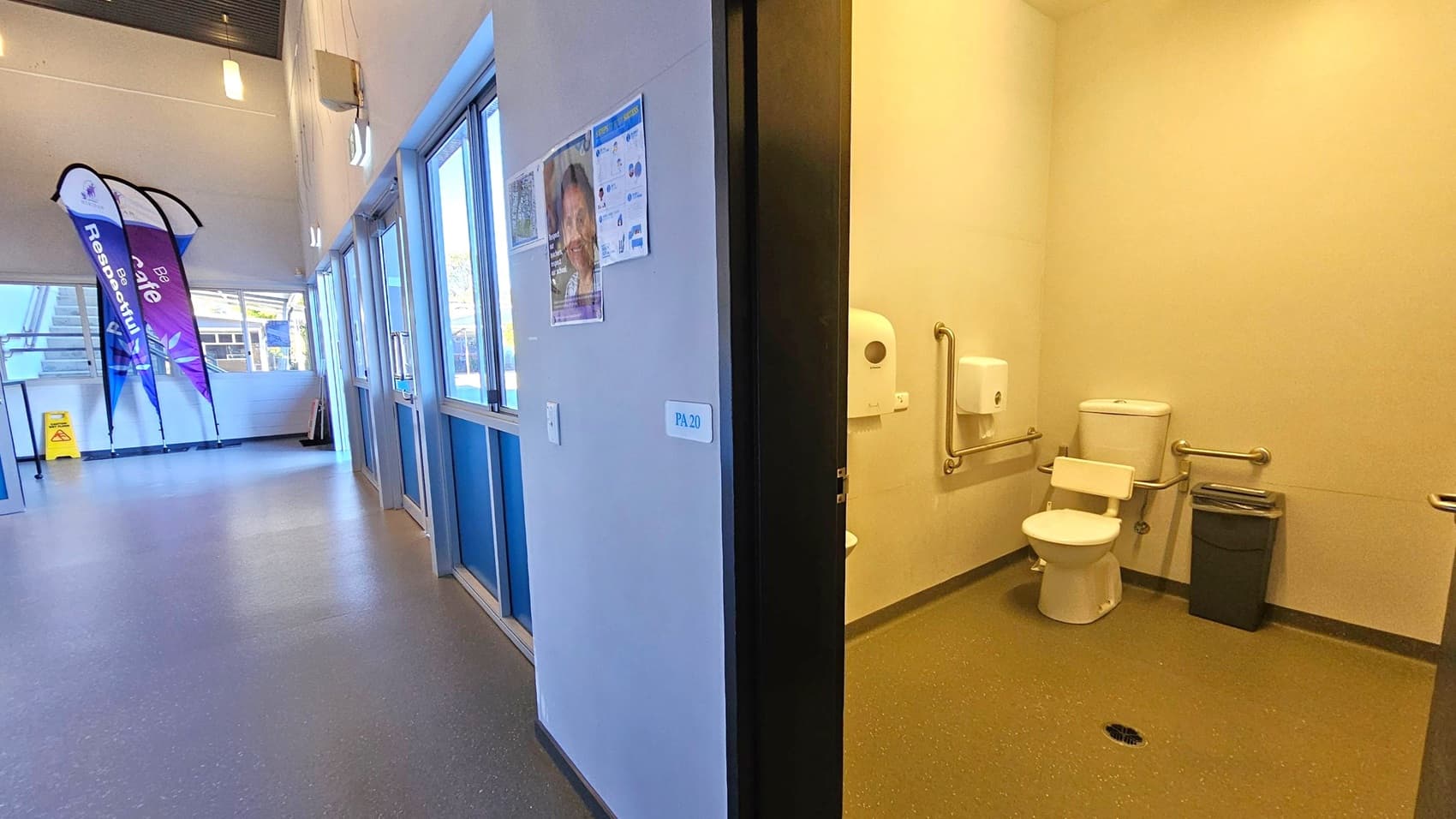 On the right, there's a bathroom with a toilet, grab bars, a paper dispenser, and a bin. On the left, a hallway with large windows, a poster on the wall, and a sign marked "PA 20". In the background, there are two vertical banners and a caution sign near a staircase. The floor is a speckled grey, and the ceiling has hanging lights.