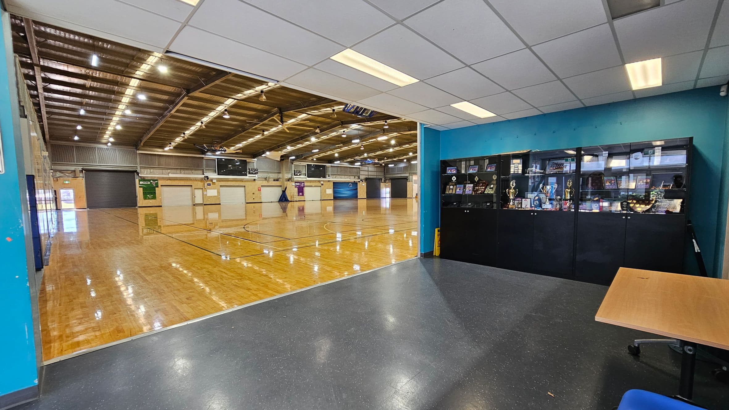 On the left, a large indoor sports hall with a polished wooden floor and high ceiling with exposed beams and lights. In the middle, the hall is open and spacious, with basketball court markings visible. On the right, a blue wall with a display cabinet filled with trophies and awards. The foreground has a dark speckled floor and a wooden table with a blue chair. The ceiling is white with fluorescent lights.