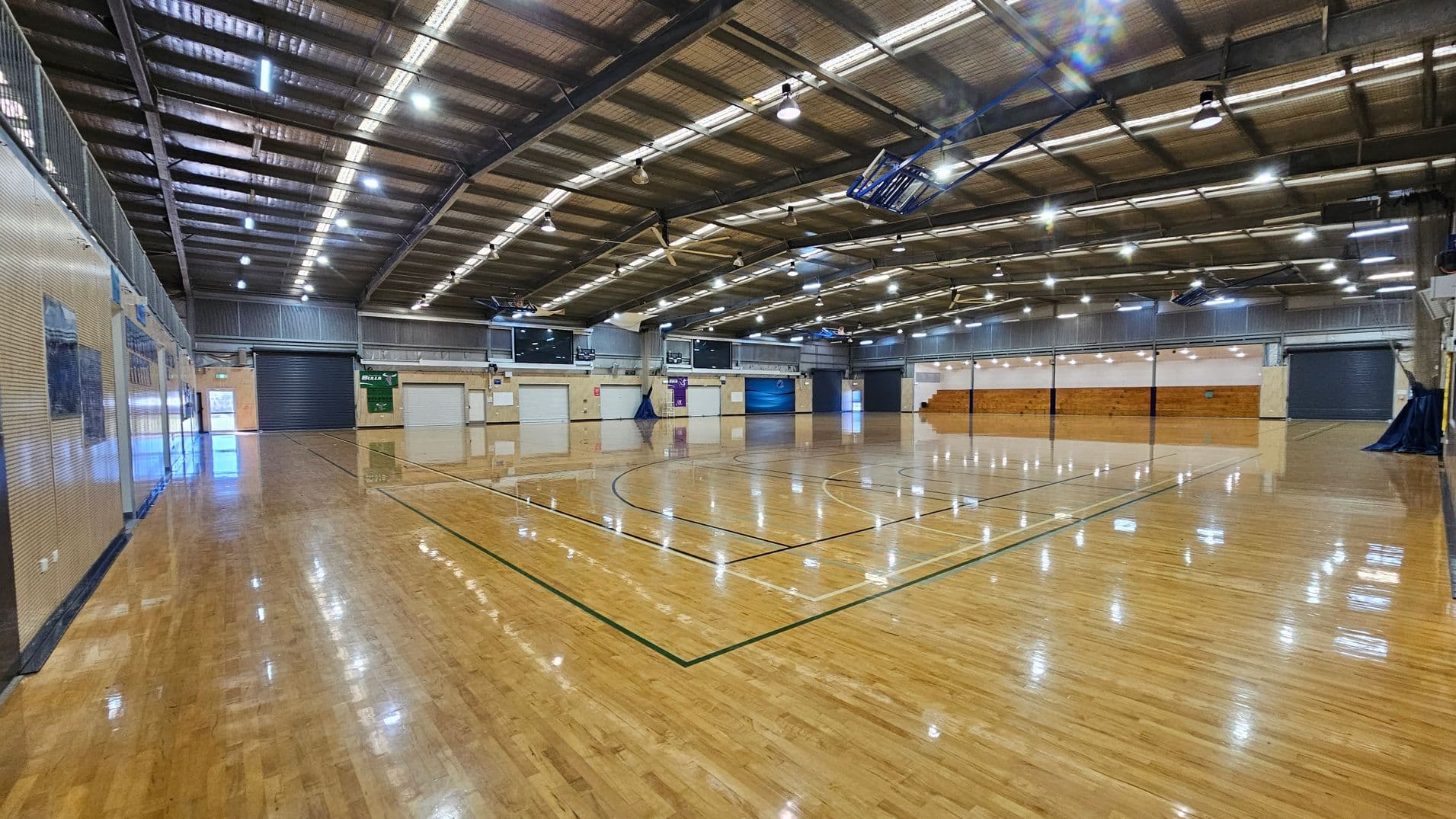 A spacious indoor sports hall with a polished wooden floor marked for basketball. The ceiling is high with exposed beams and numerous lights. On the left, there are walls with windows and doors. The right side features a large seating area with wooden benches. The background shows several closed roller doors and banners. The overall atmosphere is bright due to the overhead lighting.