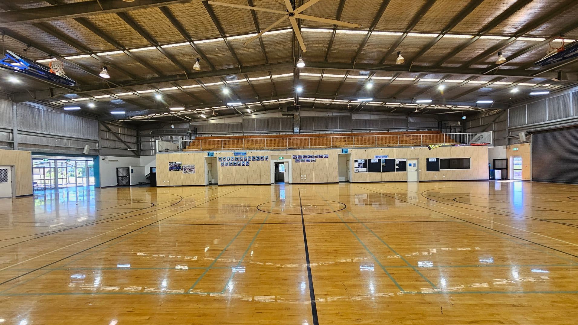 A spacious indoor sports hall with a polished wooden floor marked for basketball. The ceiling is high with exposed beams and several lights. On the left, there are large windows and doors allowing natural light. The right side features a large roller door. The back wall has a series of doors and notice boards, with some photographs and posters displayed. A basketball hoop is visible on the left side.