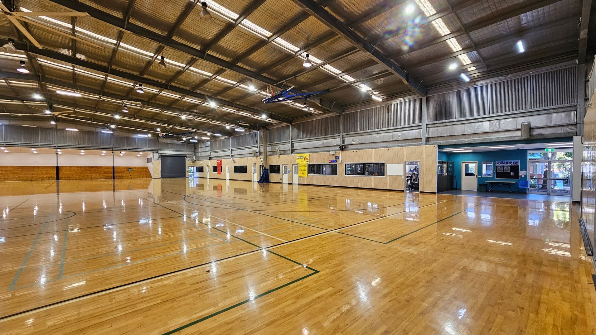 A spacious indoor sports hall with a polished wooden floor marked for basketball. On the left, there are wooden bleachers against a light-coloured wall. The right side features a wall with several windows and doors, including a blue section with a notice board and entrance. The ceiling is high with exposed beams and multiple lights. The far end has a large, closed roller door.
