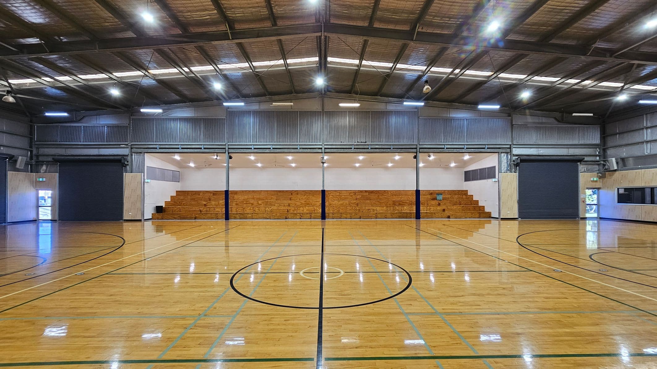 A spacious indoor sports hall with a polished wooden floor marked for various sports. In the centre, a circular marking is visible. The background features wooden bleachers for seating, flanked by two large, closed roller doors on either side. The ceiling is high with exposed metal beams and bright overhead lights. The walls are a mix of metal and wood panelling, with a small window on the right side.