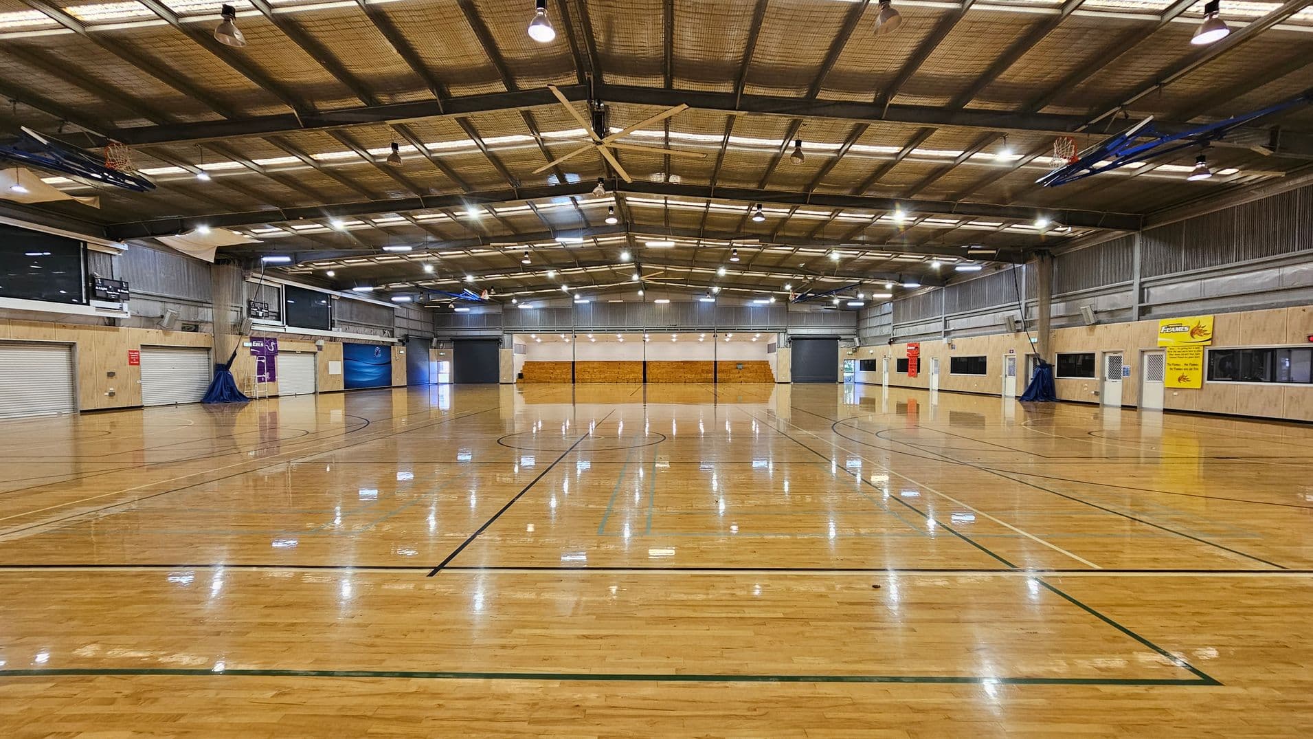 A large indoor sports hall with a polished wooden floor marked for various sports. The ceiling is high with exposed beams and multiple lights. On the left and right walls, there are several closed roller doors and windows. Banners and signs are visible on the walls. In the background, there are more roller doors and a wooden seating area. The space is well-lit and spacious, suitable for activities like basketball or volleyball.