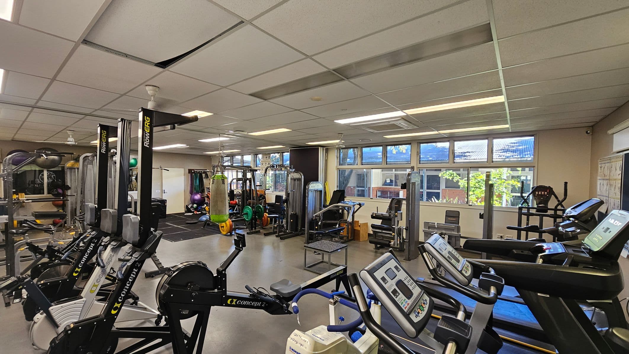 A gym with various exercise equipment. On the left, there are rowing machines and a rack with medicine balls. In the centre, a punching bag hangs from the ceiling, surrounded by weightlifting equipment and benches. On the right, treadmills and elliptical machines are positioned near large windows that let in natural light. The ceiling is made of white tiles, some of which are slightly misaligned. The floor is covered with a mix of rubber mats and hard surfaces.