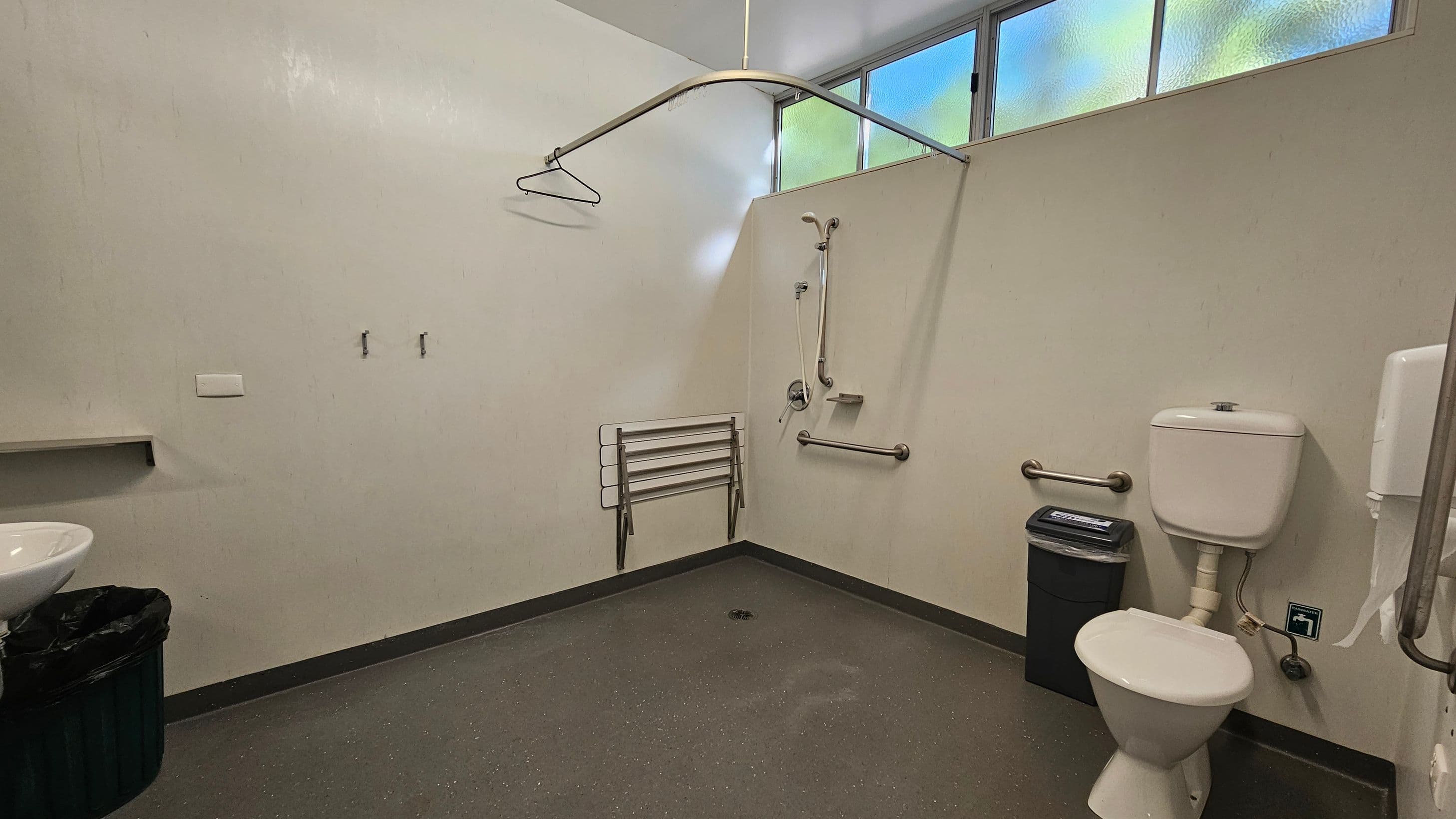 A bathroom with a white toilet on the right, equipped with a grab bar and a black bin nearby. A white sink with a black bin underneath is on the left. In the centre, a shower area features a handheld showerhead, grab bars, and a foldable shower seat. A curved shower rail with a hanger is above. The floor is speckled green, and the upper wall has frosted windows allowing light in.