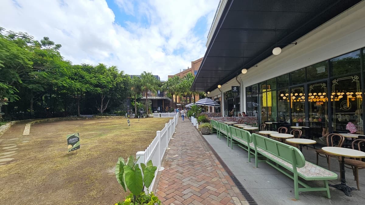To the left, a field with sparse grass and soil stretches out. Tall trees and decorative shrubs fill a concrete planter. Signs stand near a white fence. On the right, a three-story building with a brick front and large windows is visible. Large striped umbrellas line the front of the restaurant's Al Fresco area, with another brick building adjacent to a taller, multi-story structure behind it.