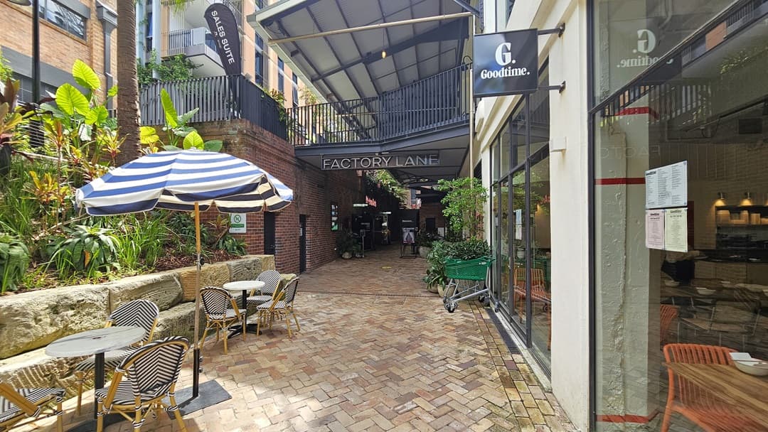 On the left, ornamental plants and palm trees line the landscape, bordered by large rocks. Nearby, a massive striped umbrella shade arranged tables and chairs. To the right, a glass wall reveals the restaurant inside the building. The brick pathway leads towards the shaded area of a red brick building. A green trolley sits near the window, accompanied by ornamental plants in large pots.