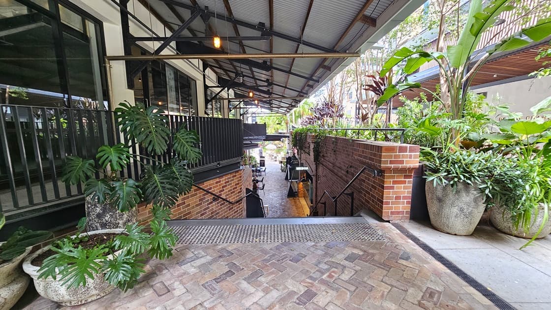 A staircase with handrails descends, preceded by truncated domes. Large pots of ornamental plants adorn the front and right sides. The left building features a corrugated roof extending to the right, secured by a low black metal fence. Brick walls flank both sides. Below, tables and chairs are visible.