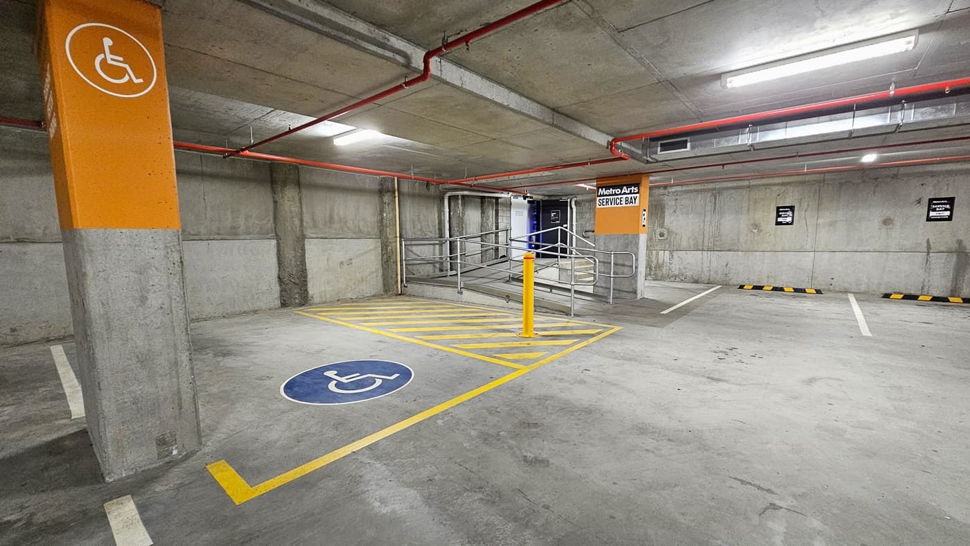 The underground car park features accessible car parking marked with yellow and blue wheelchairs, yellow diagonal lines, and a yellow bollard. A step-free ramp with handrails is on the right. The ceiling has a concrete finish with exposed orange pipes.