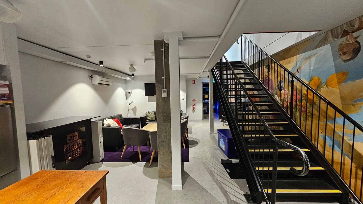 To the right, there's a staircase with handrails, under which is a blue cooler box. To the left, there's a seating area with cushioned chairs and a wooden table. More wooden tables are positioned near the wall and fridge. A large colourful painting fills the entire wall on the right. The floor, walls, and ceilings are white vinyl, equipped with LED lights.