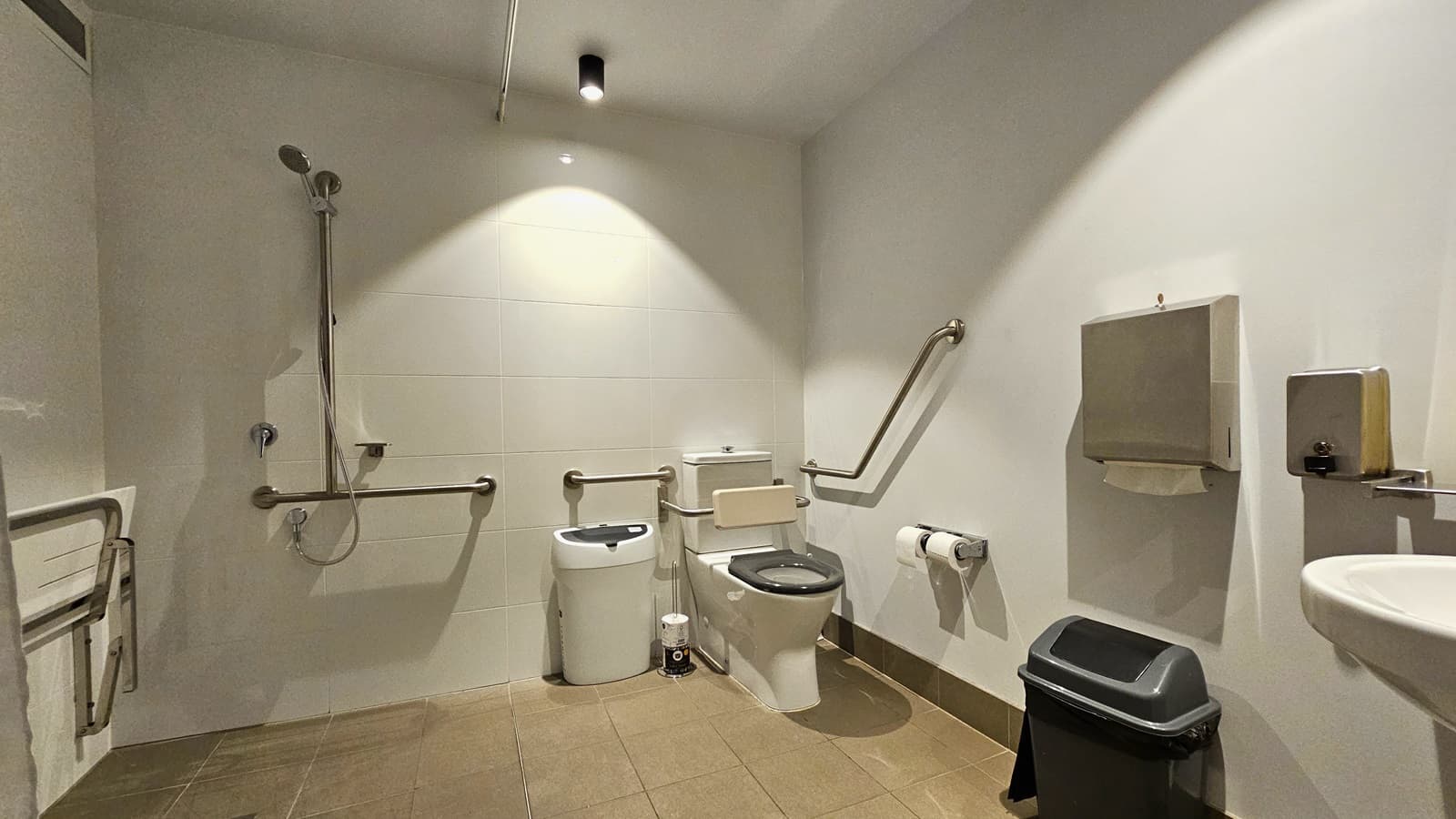 To the left, a foldable bench chair is affixed to the wall. Next to it is a shower with handrails, followed by a toilet bowl with hand grabs. A rubbish bin and tissue dispenser are beside it. To the right, there's a washbasin, another tissue dispenser, and a soap dispenser mounted on the wall. Underneath is another rubbish bin. The floor has cream tiles, while the walls and ceiling are white.