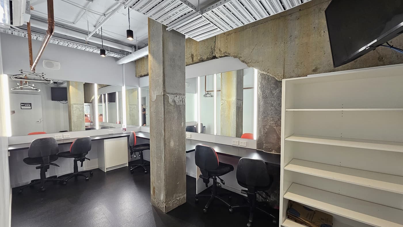 The dressing room features mirrors surrounding the walls. Adjustable chairs are positioned near the mirrors. To the right, there's a white display rack, with a flatscreen mounted on top. The ceiling displays exposed concrete beams, pipes, and air conditioning vents. The walls are white, and the floor is tiled black.