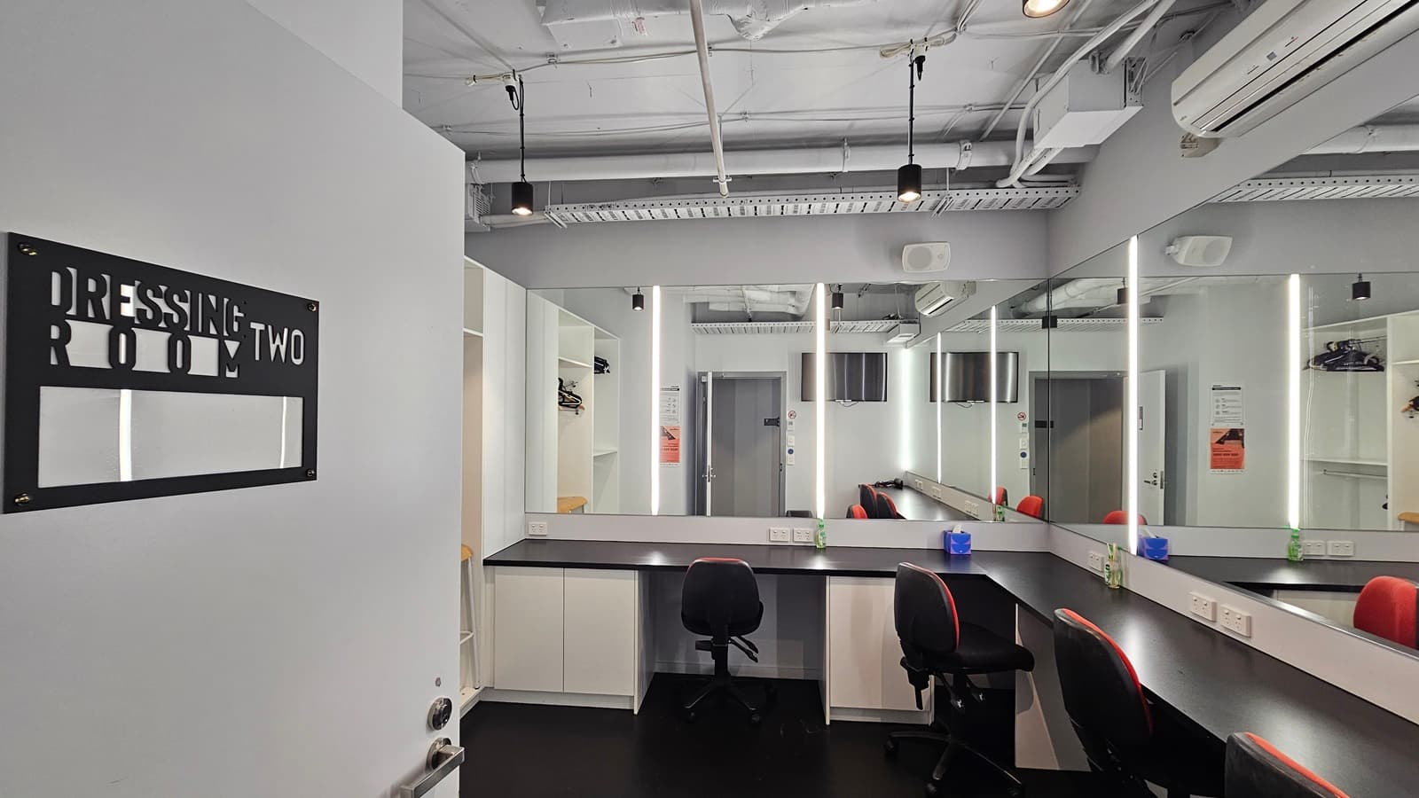 Dressing Room Two has mirrors fixed to the wall and adjustable chairs nearby. An inward-opening door with a sign is to the left. The ceiling shows exposed concrete beams, pipes, and air conditioning vents. The walls are white, and the floor is tiled black.