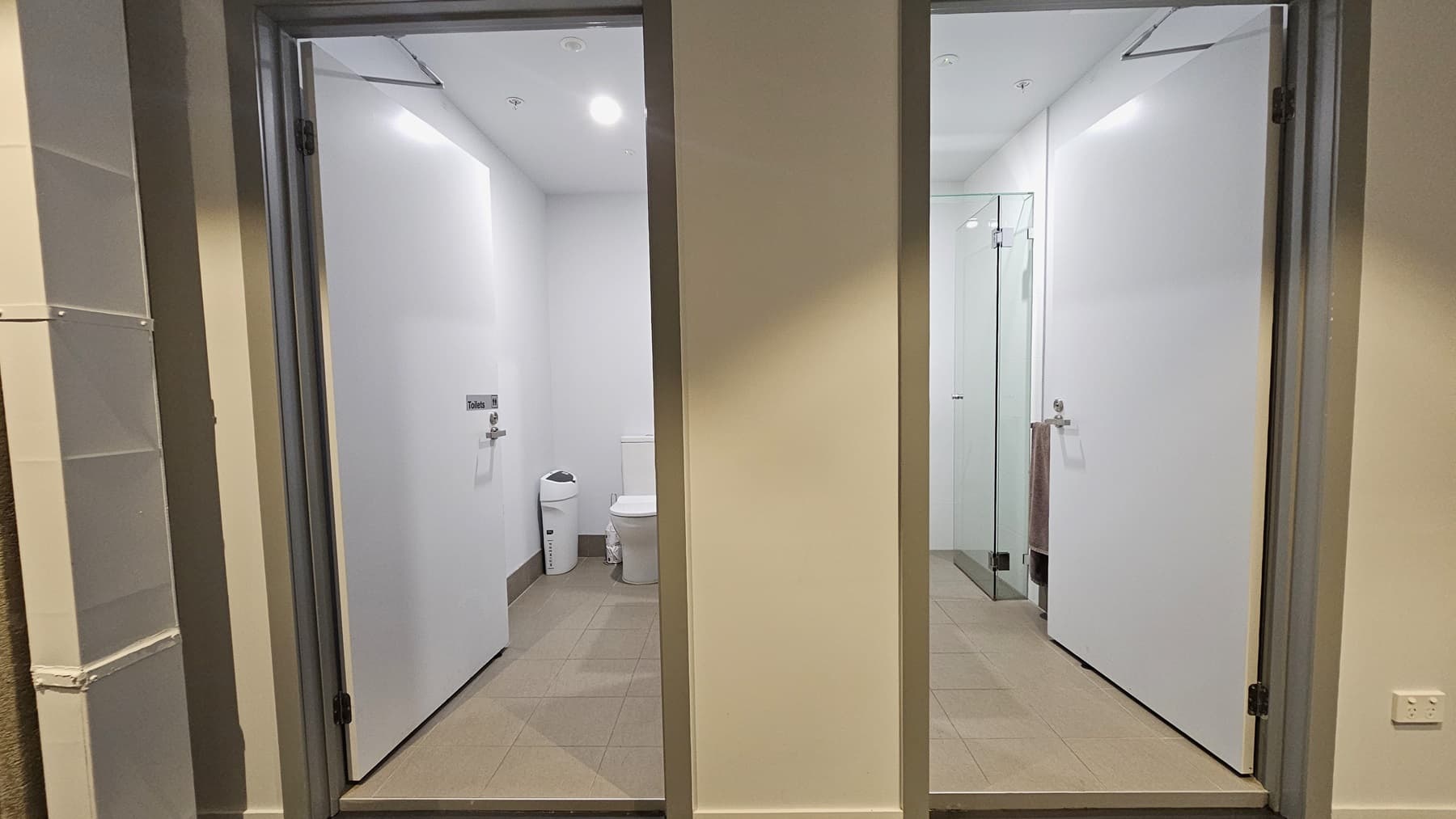 Two inward-opening doors. The first reveals a toilet bowl placed near the corner, accompanied by a white rubbish bin. The second door unveils a glass-enclosed shower area. The floor is tiled with brown, and the walls and ceiling are all white with LED lights.