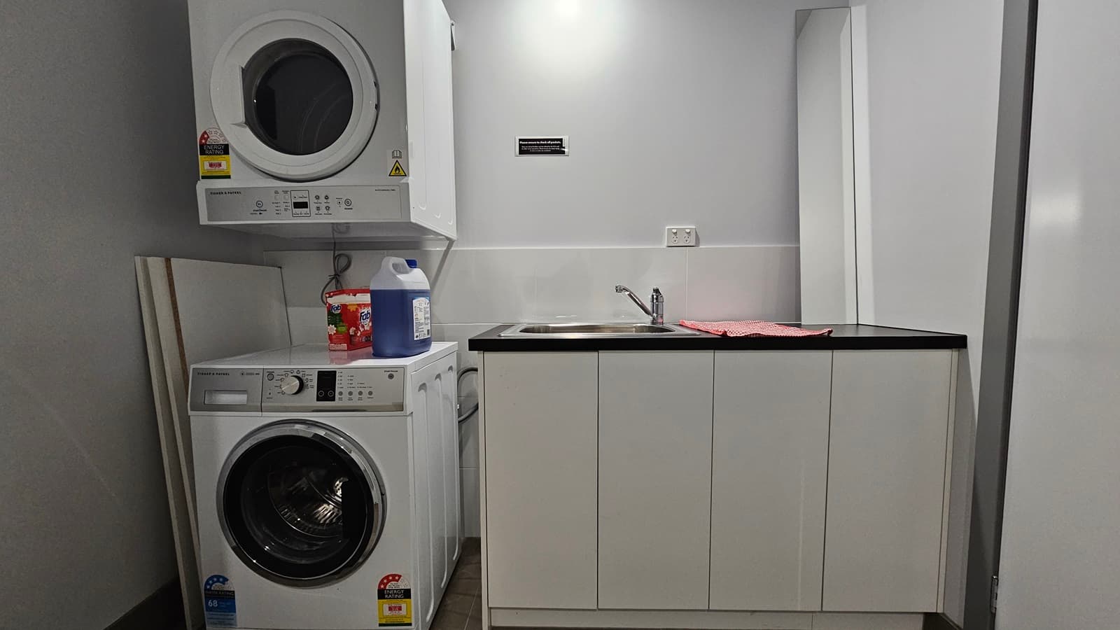 A laundry machine with detergent soaps on top. Another laundry machine is overhead. To the right, there's a small sink with a faucet, and a kitchen towel placed on top. The walls are all white, and the floor has brown tiles. An instruction sign is placed on the wall.
