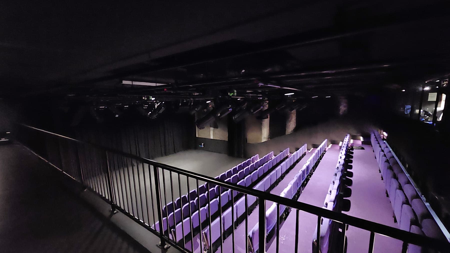 A dimly lit area with black floor tiles and walls. Metal railing secures the space. Overlooking below is the stage with black tiles, surrounded by black walls and curtains. Adjacent to the stage is tiered seating with lavender chairs. The ceiling features exposed pipes, audio equipment, and lighting fixtures.