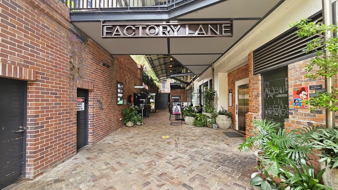 Both sides of the building display a red brick facade with black doors. A ceiling-hung signage reads "Factory Lane". On the right, a lift is positioned. Large pots showcase ornamental plants upfront, alongside free standing banners. Posters adorn the brick walls, with a concrete staircase visible further ahead.