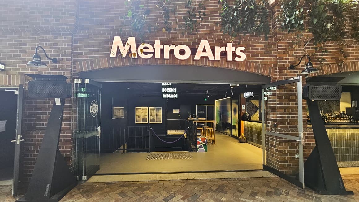 On the left stands a building with a brick facade labelled "Metro Arts". Ornamental plants hang above, extending towards the building. Lighting fixtures adorn the wall, with two black speaker-like structures positioned in front of the glass door entrance. Inside, a staircase with handrails is equipped with truncated domes and a rope bollard. To the right, a bar is arranged with movable chairs for seating.