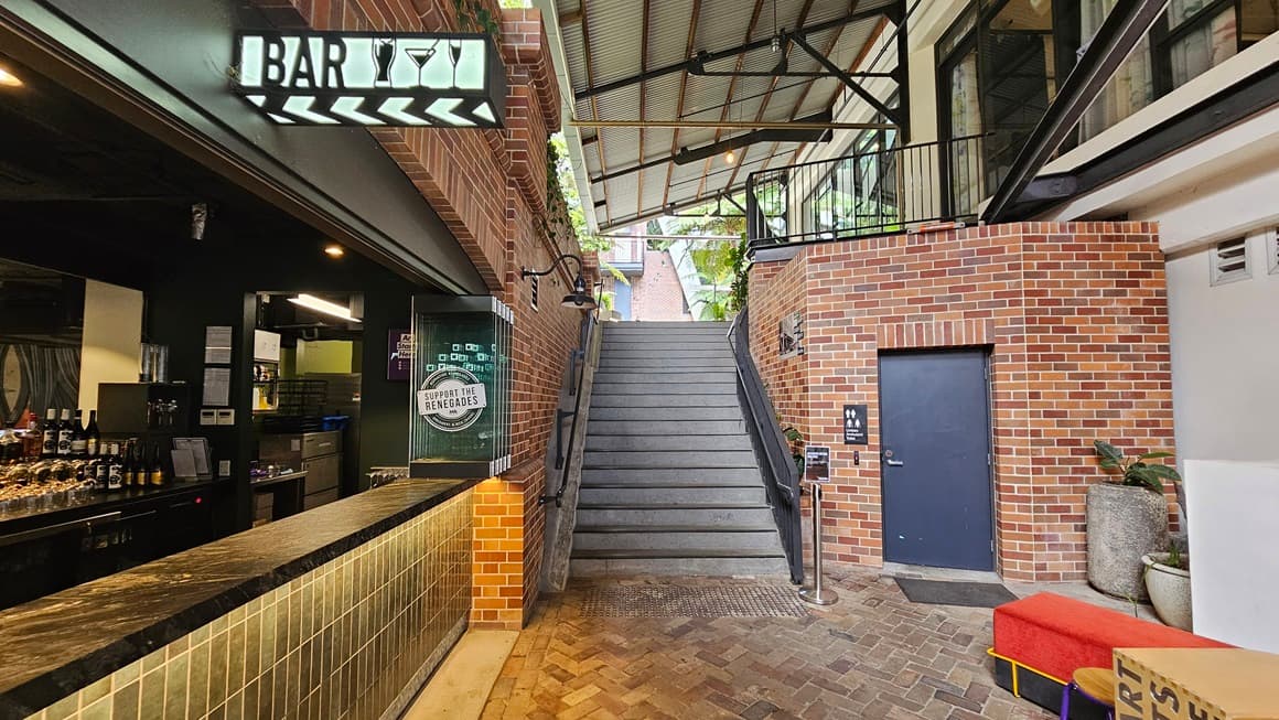 To the left stands a building with a brick facade, with a neon sign reading "Bar". Inside, a brick wall decorates the bar area, with wines and liquors displayed on the counter. A staircase equipped with handrails and truncated domes is positioned in the middle. To the right, an accessible toilet with a brick facade and grey door is situated. Ornamental plants in large pots adorn the right side, while the corrugated roof extends to the left, providing shade. Brick tiles cover the floor.