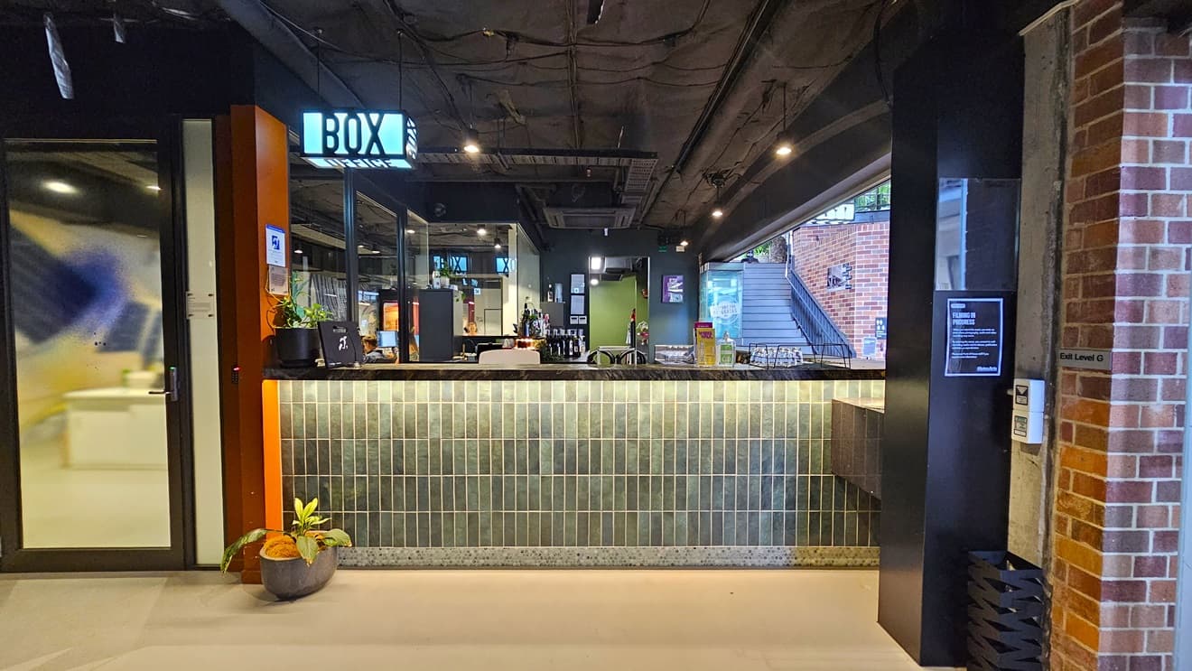 The bar showcases a brick facade with a neon sign reading "Box Office". Wines and liquors are displayed on the counter. A glass door and a pot of ornamental plants adorn the left side. A staircase with handrails and truncated domes is positioned to the right. Signages are affixed to the wall. The ceiling is all black with lights and aircon vents, while the floor is tiled white.
