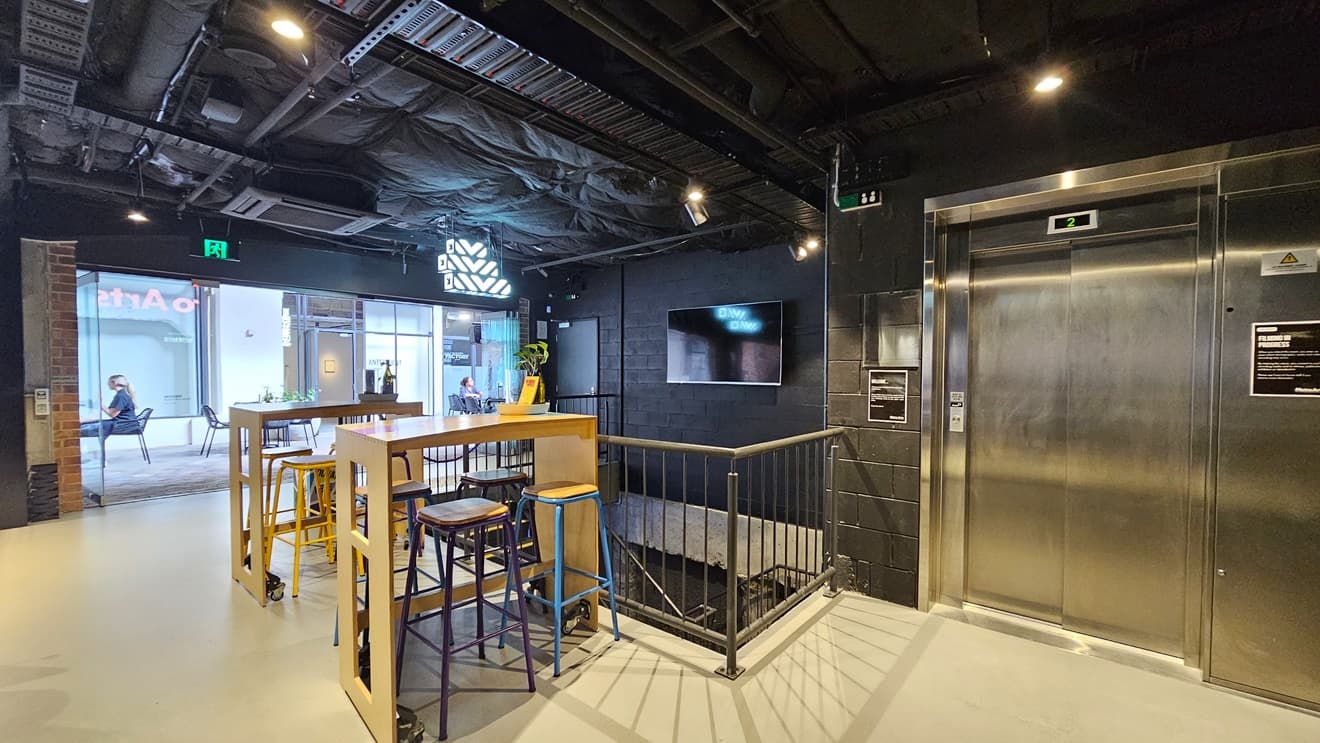 To the right, a lift displays a poster on its wall, adjacent to a flatscreen attached to the black wall. In the middle, two tables with stools are situated, while a staircase leading downwards stands behind. Metal railing secures the area from the outside. The ceiling is all black with lights and aircon vents, and the floor is tiled white.