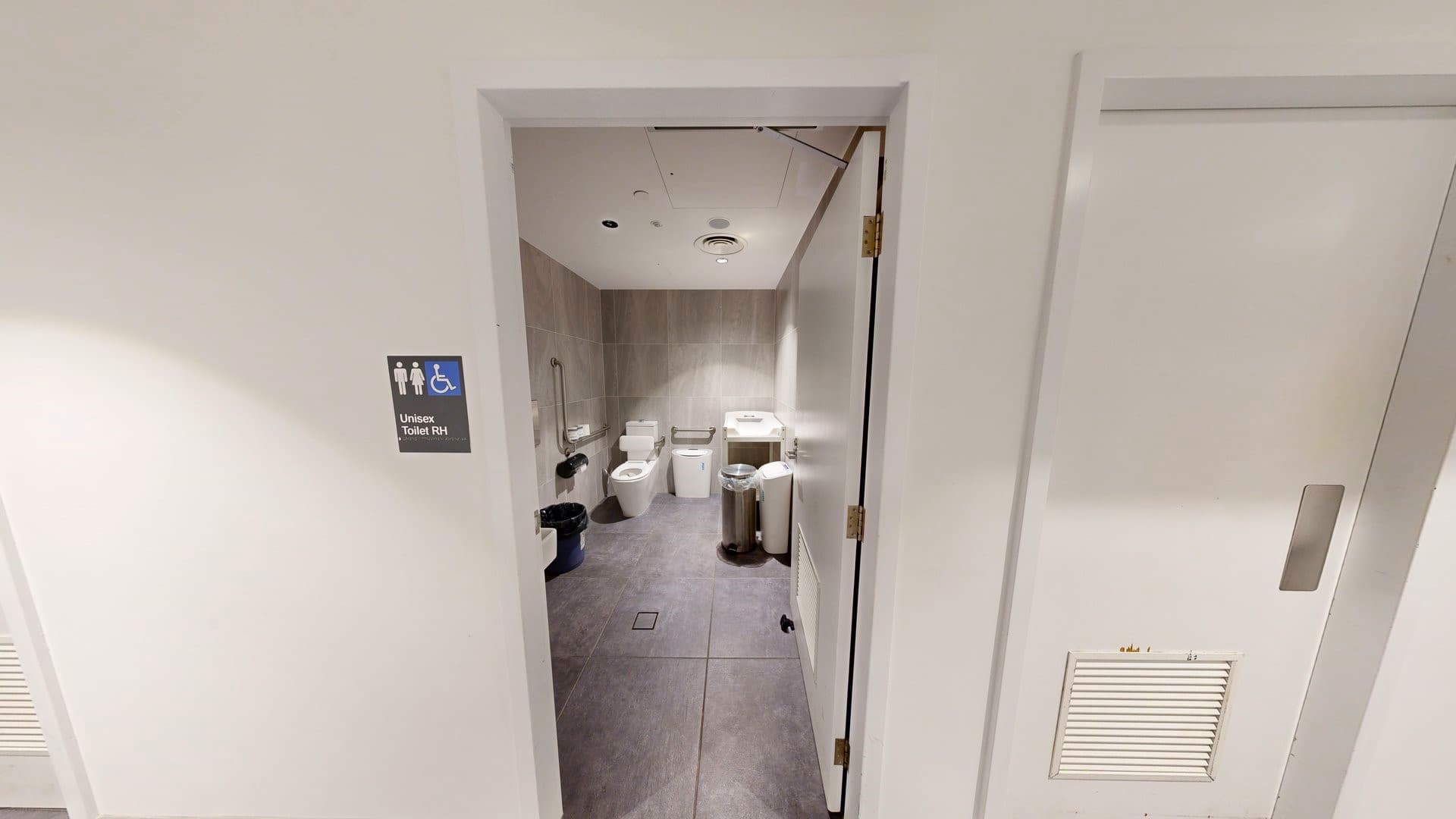 In the unisex bathroom, a toilet bowl is in the left corner with handrails and a tissue dispenser on the wall. A washbasin is positioned to the right alongside rubbish bins. Grey tiles cover the floor and walls. The white ceiling is illuminated by LED lights. Outside, signage indicates all-gender access on the white wall next to another white door.