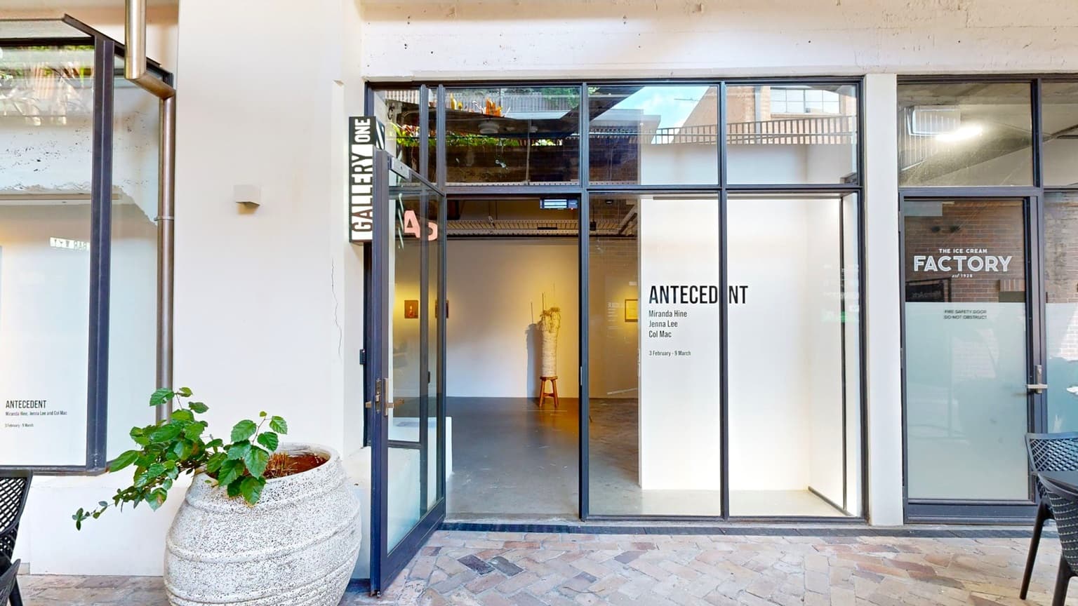 A manual glass door opens outward, with a signage reading "Gallery One" on the wall above. An ornamental plant sits in a huge pot below. Signages are affixed to the right side of the glass door. Movable black tables and chairs are arranged in front. The floor is tiled with brick, and the wall is white. Inside, a section of the gallery is visible.