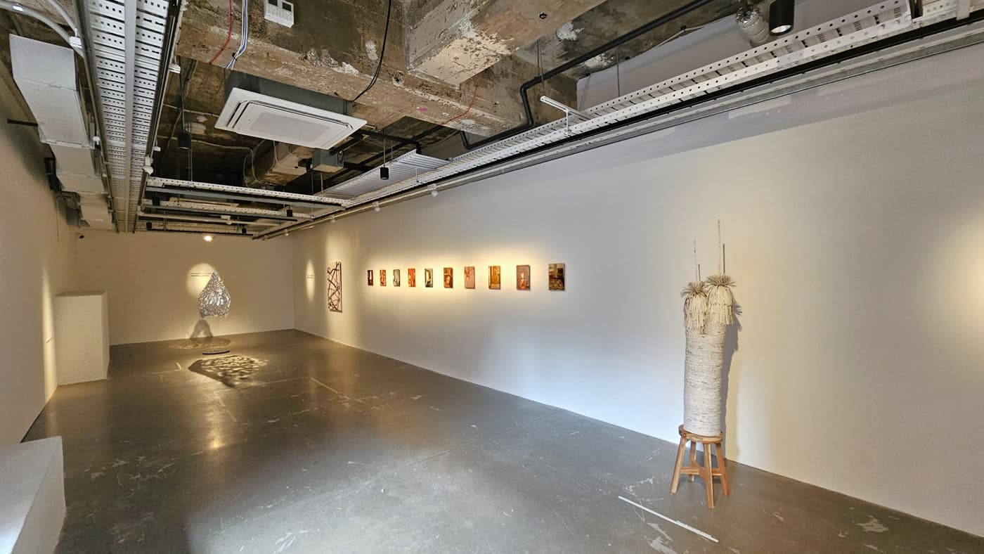 To the right, art paintings adorn the white wall, accompanied by hanging lighting fixtures. A piece of craft made of ropes and strings is displayed against the wall. The ceiling reveals exposed concrete beams, pipes, air conditioning vents, and pin lights. The polished cement floor exhibits worn-out markings.