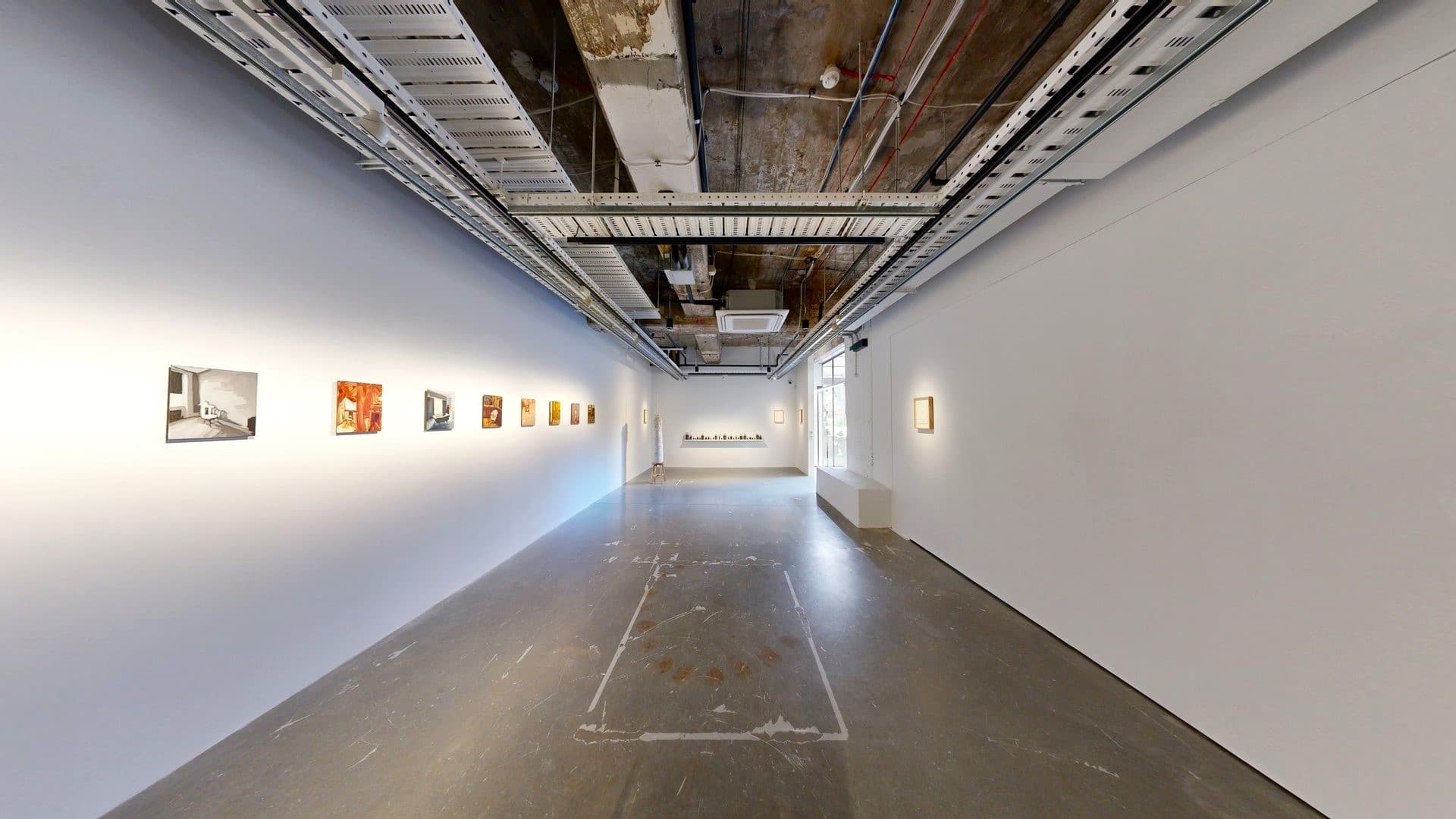 To the left, art paintings decorate the white wall, with another piece on the right. A craft made of ropes and strings is displayed against the far-left wall. The ceiling exposed concrete beams, pipes, air conditioning vents, and pin lights. The polished cement floor displays worn-out markings.