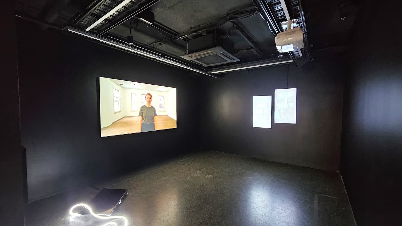 dimly lit room with black tiles, walls, and ceiling. Exposed pipes, a projector, and lighting fixtures adorn the ceiling. A large flatscreen TV is mounted on the left wall.