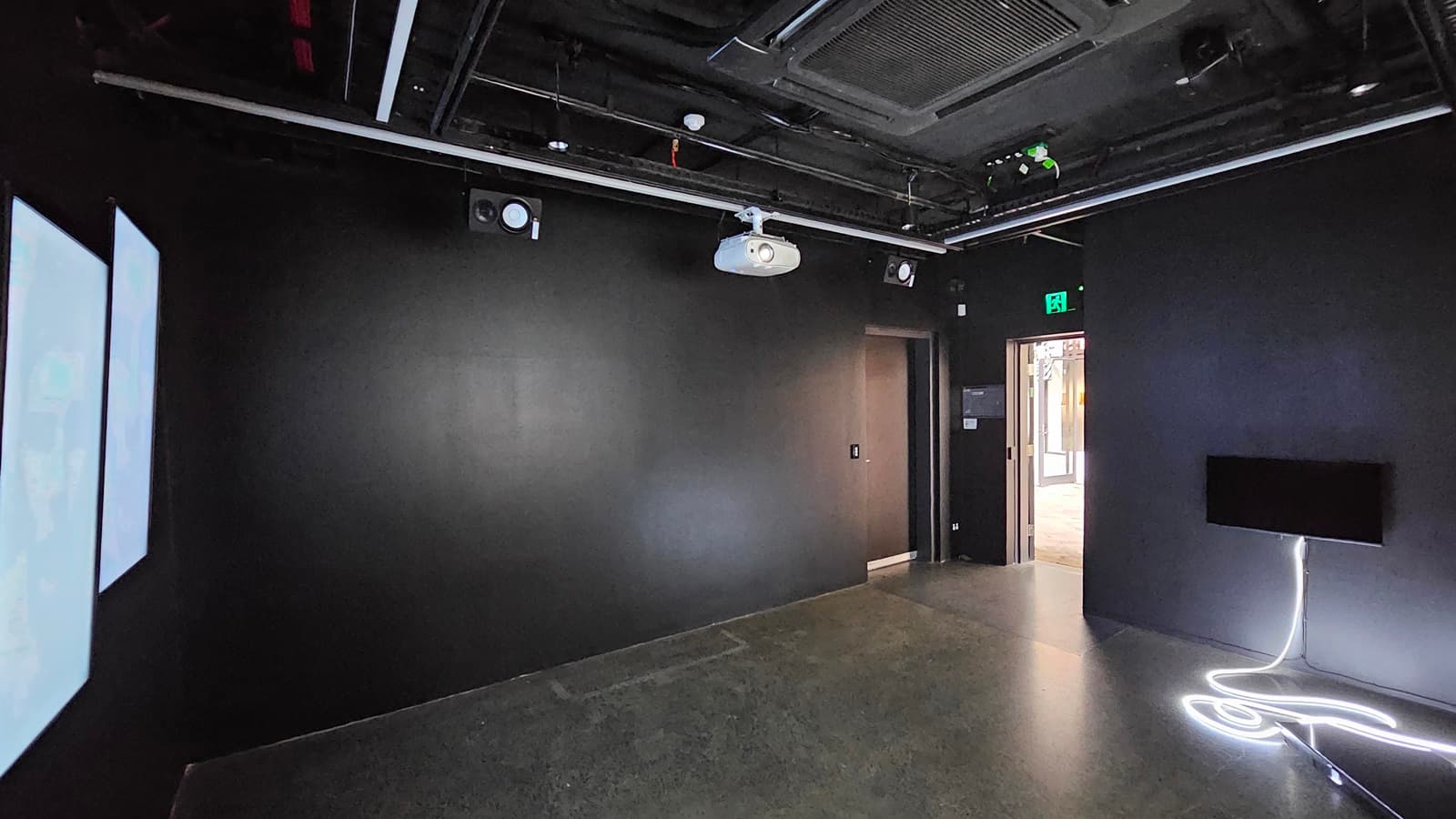 A dimly lit room with black tiles, walls, and ceiling. Exposed pipes, a projector, and lighting fixtures are visible on the ceiling. Digital screens are mounted on the left wall, while a flatscreen is posted to the right. A black door is positioned in the right corner.