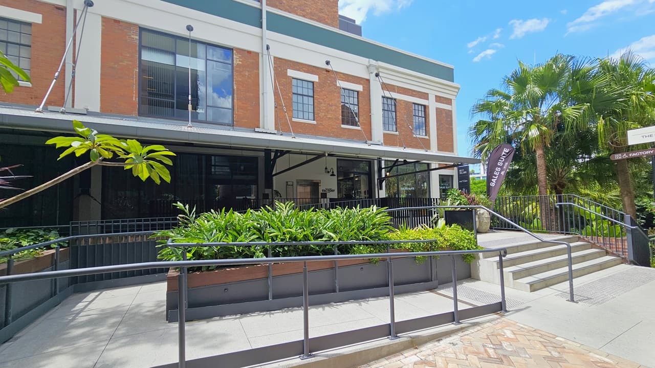 A staircase and ramp with handrails provide access to a two-storey brick building, with truncated domes installed before them. Ornamental plants adorn the concrete planter box in front. Palm trees and additional trees and ornamental plants are situated to the right. Signages are placed on the building's corner.