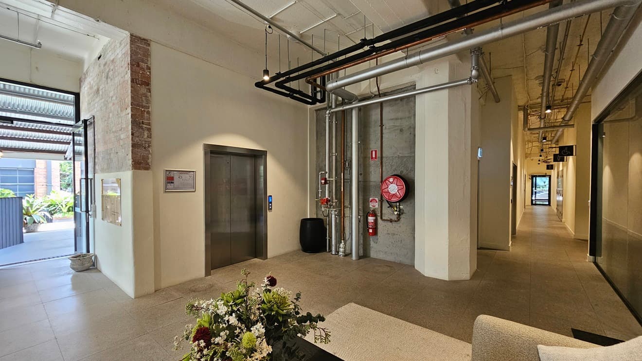 To the left, there's an inward-opening glass door with a small pot nearby. Adjacent is a lift displaying a site map poster. On the concrete wall, a fire extinguisher, fire hose reel, and exposed pipes are mounted. Below is a black rubbish bin. To the right, there's a seating area with a table holding a flower vase. Further in, a corridor leads. The bare ceiling features large pipes, air conditioning vents, and LED lights.