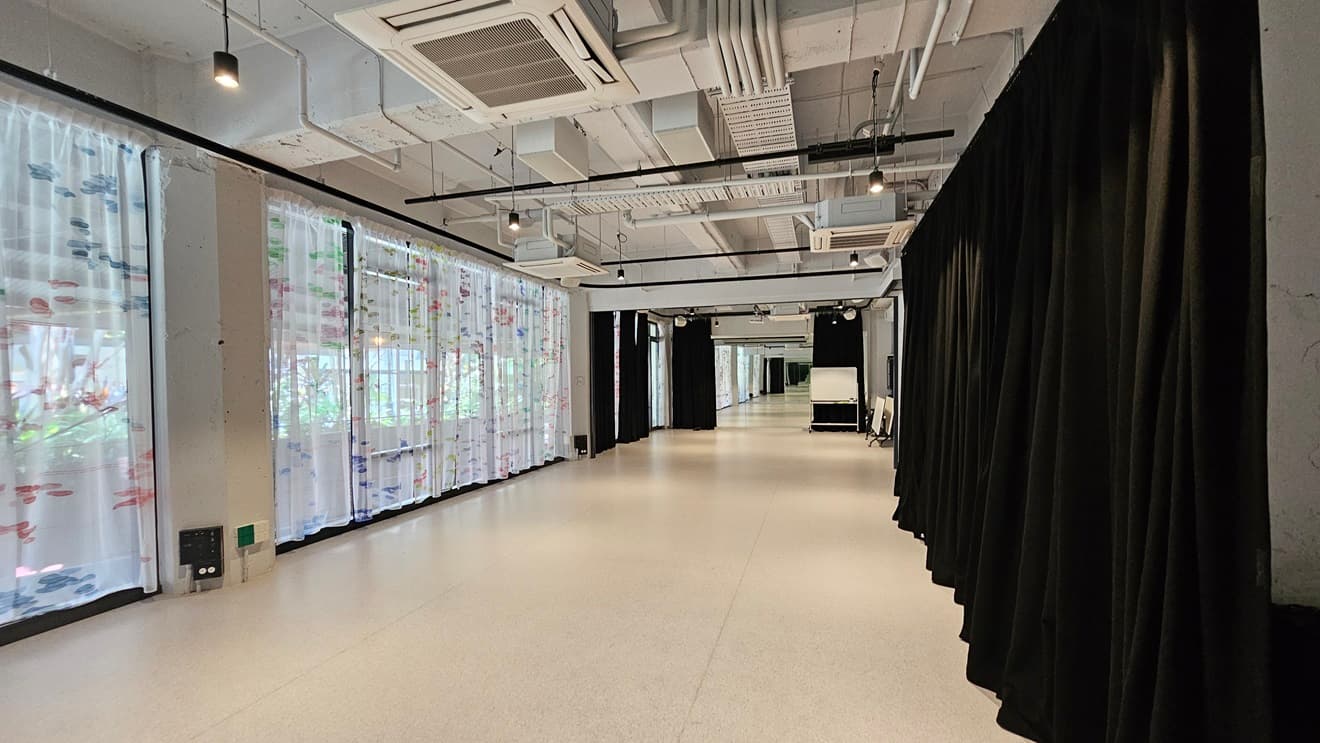 A winding space with white vinyl flooring. To the left, a floor-to-ceiling glass wall with printed white curtains; to the right, black curtains. The ceiling has an industrial look with exposed pipes, air conditioning vents, and LED lights. Foldable tables are visible on the right.