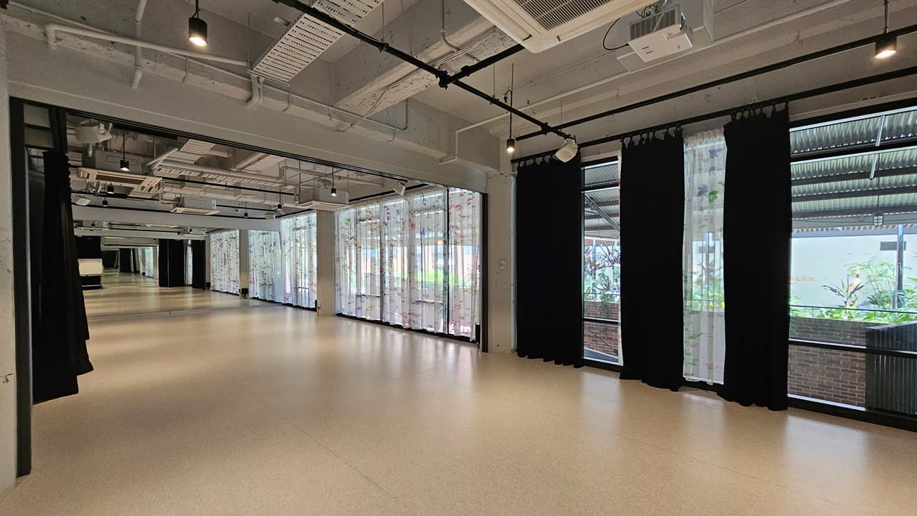 A winding space with white vinyl flooring. To the right, a floor-to-ceiling glass wall with printed white curtains and black curtains. To the left, black curtains hang from the ceiling. The ceiling has an industrial look with exposed pipes, air conditioning vents, and LED lights.