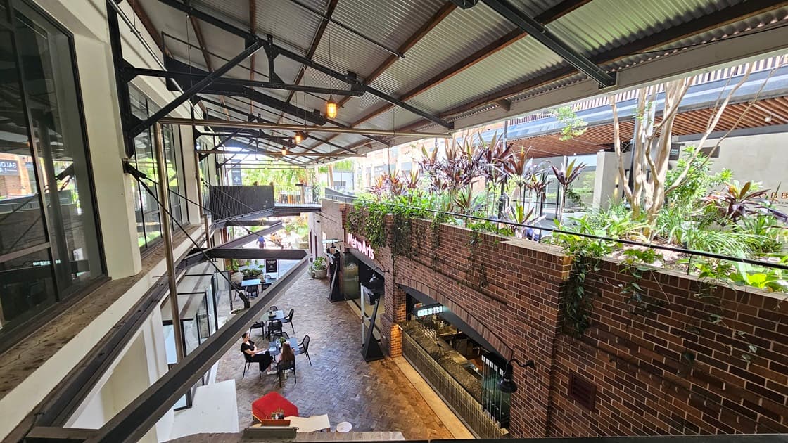 The extended corrugated roofing is supported by large metal beams, with light fixtures hanging from the top. To the right, a landscape garden features ornamental plants hanging from the brick wall. Below, The Metro Arts Bar is visible, with occupied tables and chairs.