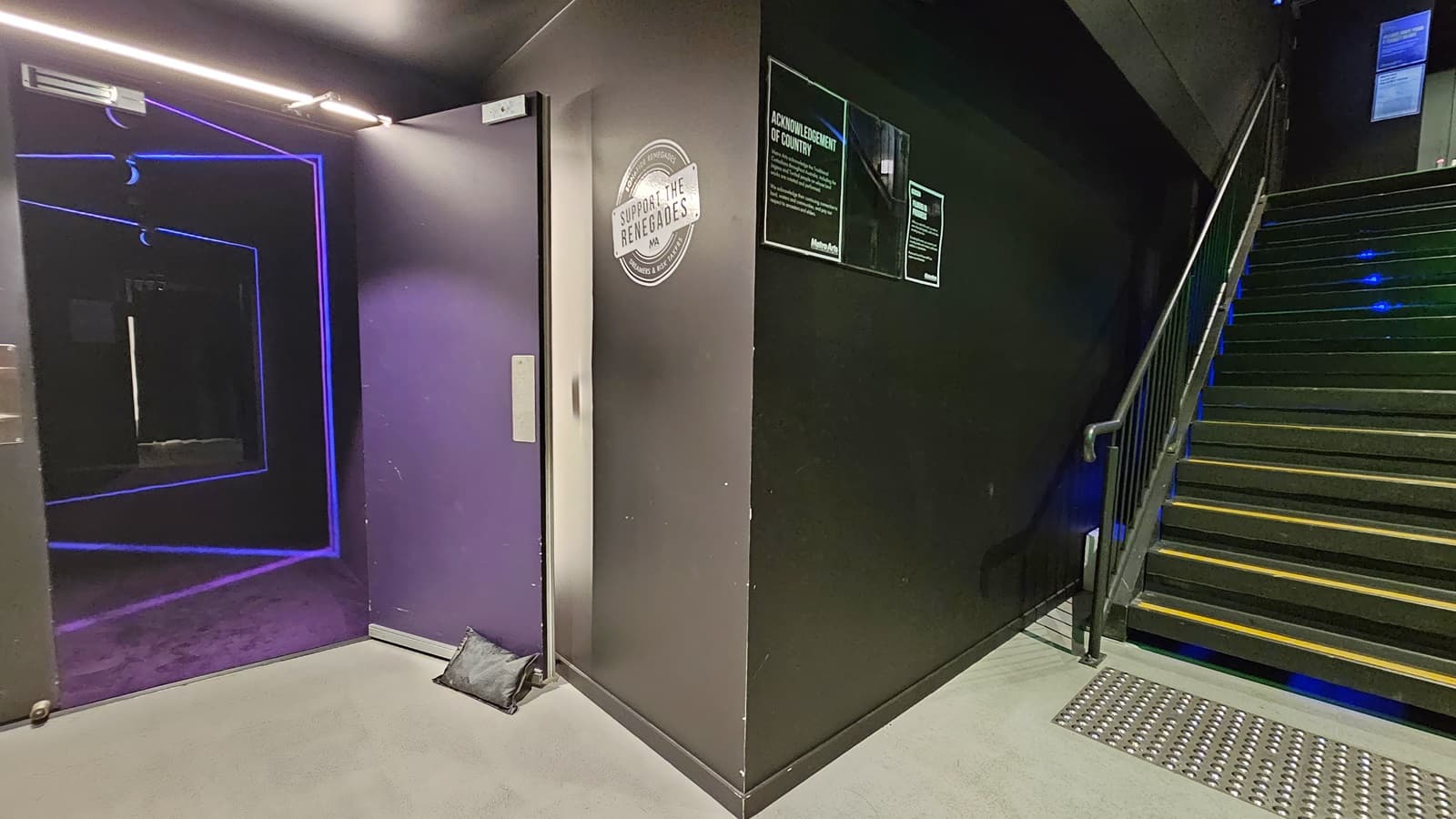 A dimly lit area with black tiles and walls. On the left, a staircase equipped with handrails and truncated domes. The right wall features glowing directional arrows pointing down. The left wall has a rough finish. Further down the stairs, a fire hose reel is visible.