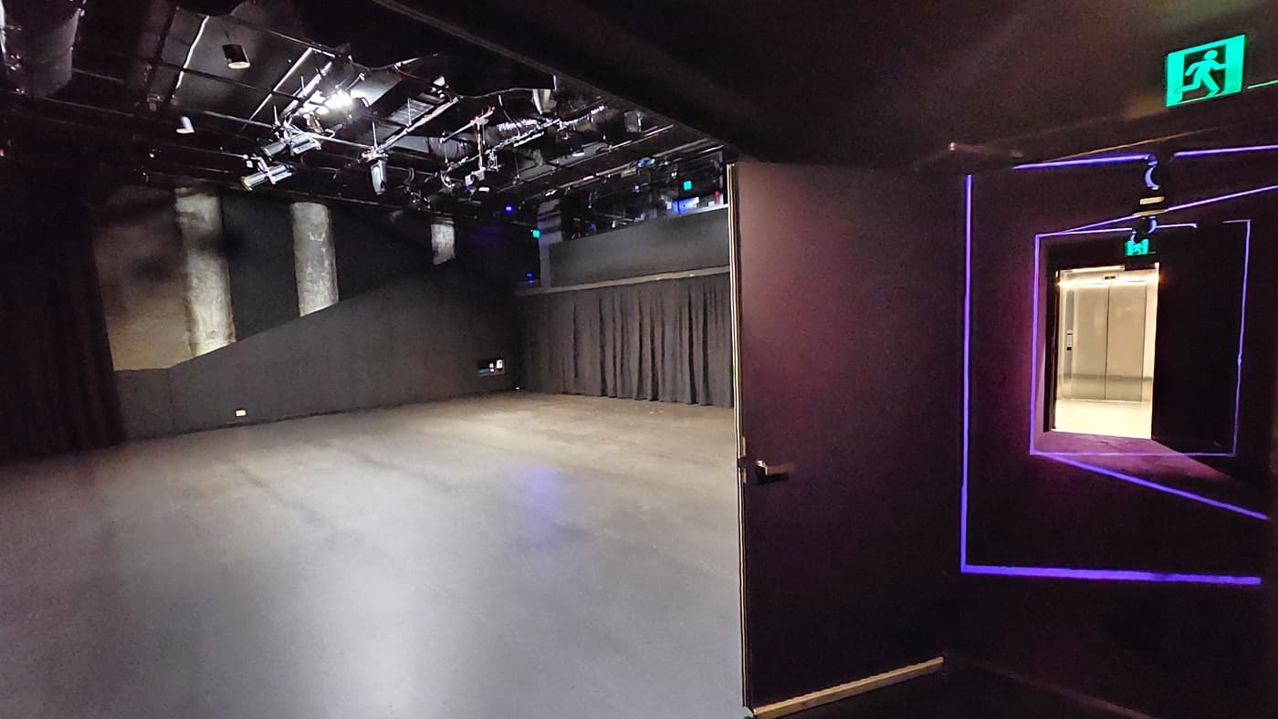 In a dimly lit space with black floor tiles and walls, an exit door to the right is illuminated by neon lights, leading to the lift. To the left, a stage with black tiles and curtains is visible. The ceiling is covered with black insulation, housing audio, and light fixtures.