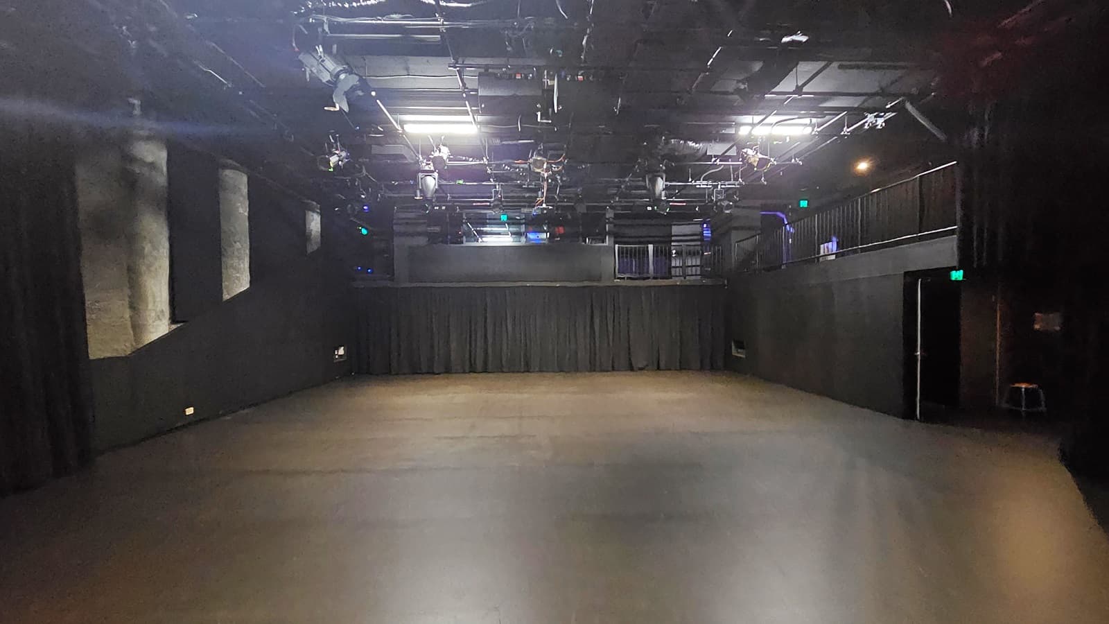 In a dimly lit space with black floor tiles and walls, a door to the right is illuminated by a neon sign reading "Exit". The stage is equipped with black tiles and black curtains. The ceiling is covered with black insulation, hosting audio and light fixtures. To the right, a mezzanine is secured by black metal railings.