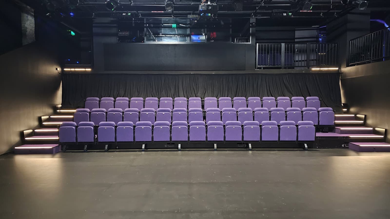 In a dimly lit space with black floor tiles and walls, tiered seating is equipped with lavender chairs. Stairs on both sides lead to the upper tier. The ceiling is covered with black insulation, housing audio, and light fixtures. Behind the seating, a mezzanine secured by black metal railings houses the controls.