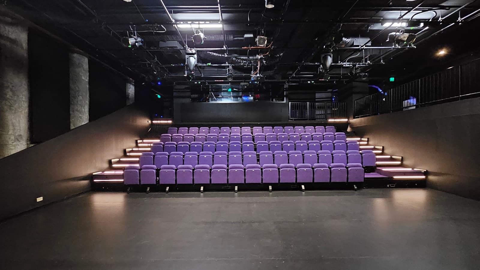In a dimly lit space with black floor tiles and walls, tiered seating is equipped with lavender chairs. Stairs on both sides lead to the upper tier. The ceiling is covered with black insulation, housing audio, and light fixtures. Behind the seating, a mezzanine secured by black metal railings houses the controls.