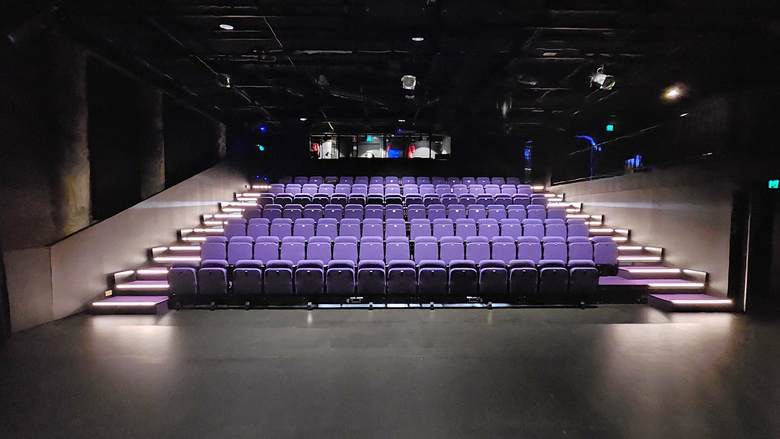 In a dimly lit space with black floor tiles and walls, tiered seating is equipped with lavender chairs. Stairs on both sides lead to the upper tier. The ceiling is covered with black insulation, housing audio, and light fixtures. Behind the seating, a mezzanine secured by black metal railings houses the controls