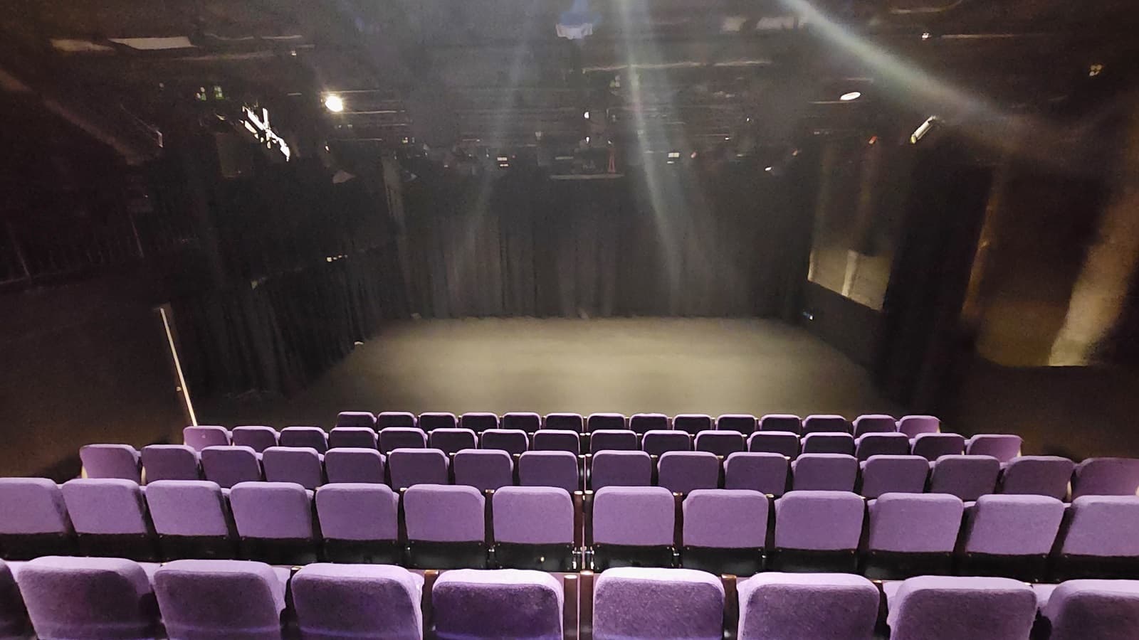 In a dimly lit space with black floor tiles and walls, tiered seating features lavender chairs. The stage in front is adorned with black tiles and surrounded by black walls and curtains.