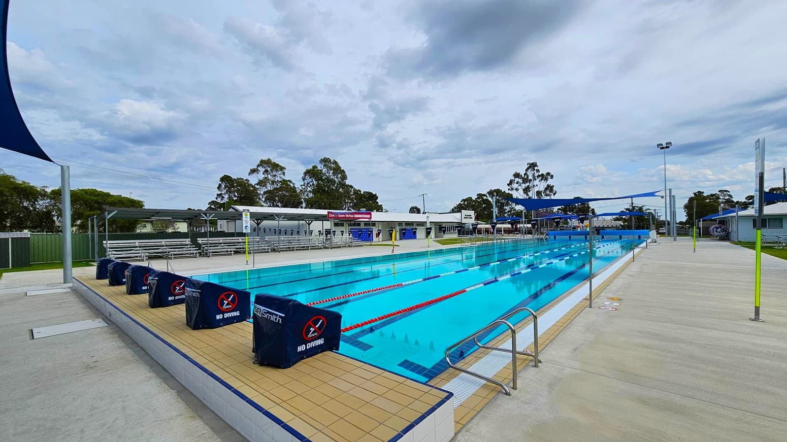 An outdoor swimming pool with clear blue water, divided into lanes by red and white ropes. On the left, there are covered spectator stands with white benches. The pool deck is tiled, with "No Diving" signs on the pool's edge. On the right, a wide concrete walkway runs alongside the pool, with blue shade sails overhead. In the background, there are trees and a low building with signage.