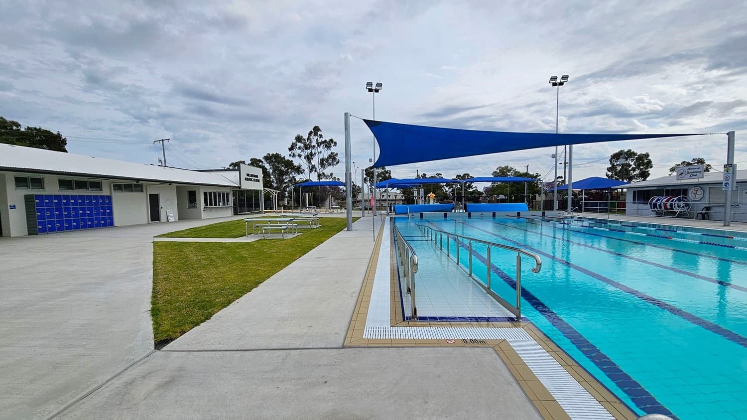Outdoor swimming pool with clear blue water on the right, featuring lane dividers and a metal handrail. Blue shade sails are stretched above the pool. On the left, a building with blue lockers and a sign reading "Aquatic Centre" is visible. There is a grassy area with picnic tables between the building and the pool. The ground is paved with concrete, and there are trees in the background.