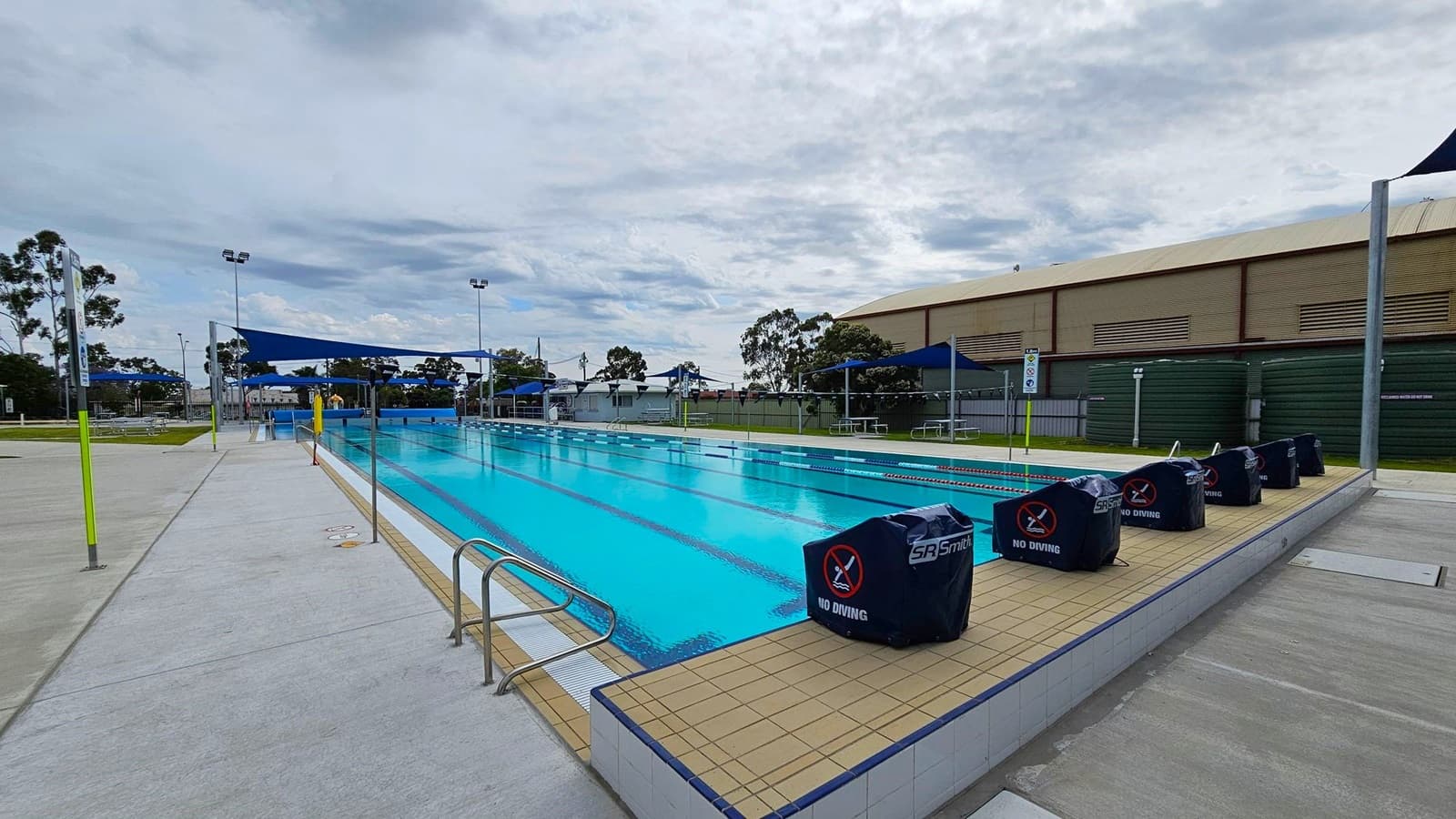 An outdoor swimming pool with clear blue water, surrounded by a tiled deck. On the right, there are covered starting blocks with "No Diving" signs. The left side features a concrete walkway with a yellow pole. In the background, there are blue shade structures, picnic tables, and a large green building. Trees and light poles are visible in the distance.