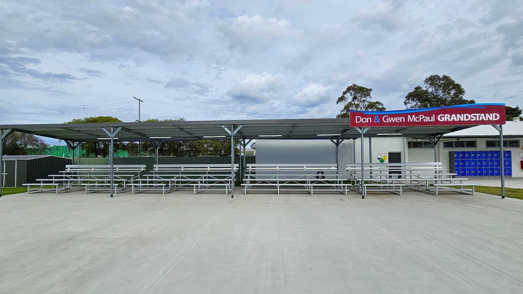 A grandstand with a metal roof and several rows of metal bleachers. The sign reads "Don & Gwen McPaul Grandstand" in red and white. Behind the grandstand, there are trees and a building with blue lockers and a door. The ground is paved with concrete, and there is grass visible on the left side.